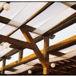 A detailed view of a rustic wooden pergola with white fabric and round light bulbs.