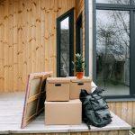 Wooden porch with cardboard boxes and potted plant next to glass windows.